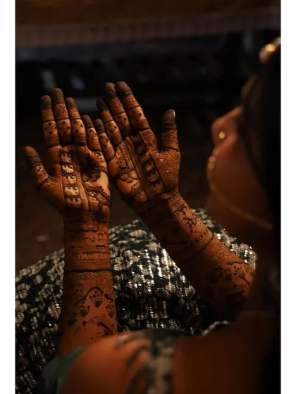 The beauty of a fresh henna stain in a dimly lit, atmospheric setting. This over-the-shoulder view highlights the complexity of the patterns on the bride's hands.