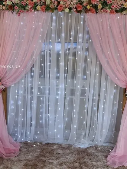 This elegant backdrop shows that I work with more than just balloons. I used soft pink and white drapes, a wall of fairy lights, and artificial flower arrangements to create a sophisticated photo corner for a home event.