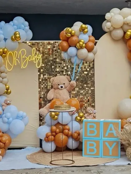 This is a popular teddy bear theme for a baby shower. The setup includes a gold shimmer wall, an 'Oh Baby' neon sign, and balloon arches in soft brown, beige, and blue. I also provide props like the 'BABY' blocks and a cake stand.